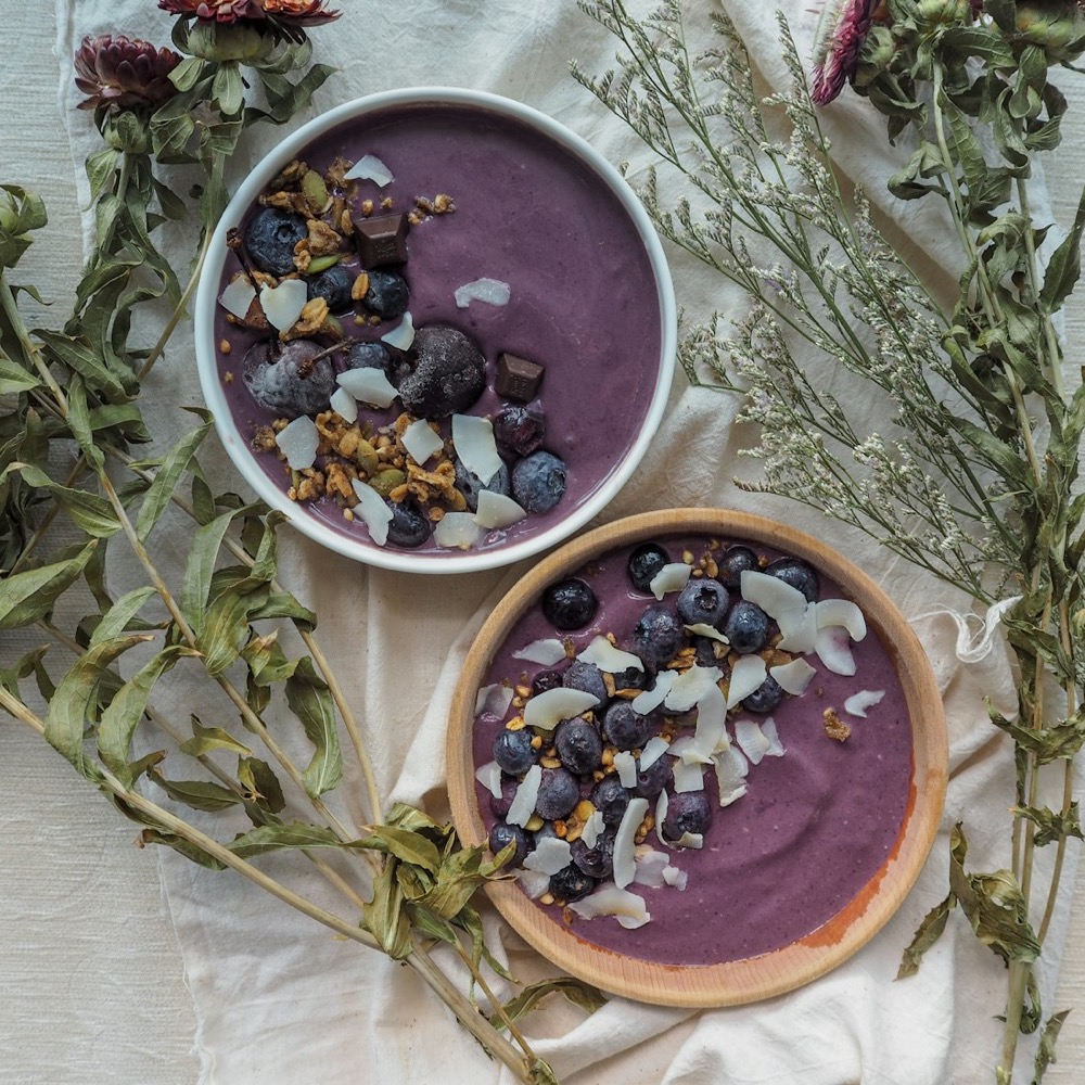 Two açaí bowls topped with blueberries, coconut and granola, styled with dried botanicals on linen